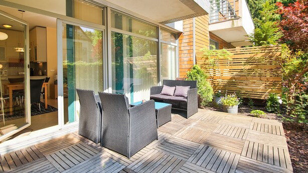 A modern outdoor patio with wicker furniture, a wooden deck, and a glass door leading to a kitchen area. - Apartamenty Swinoujscie - Marinos (Swinoujscie)
