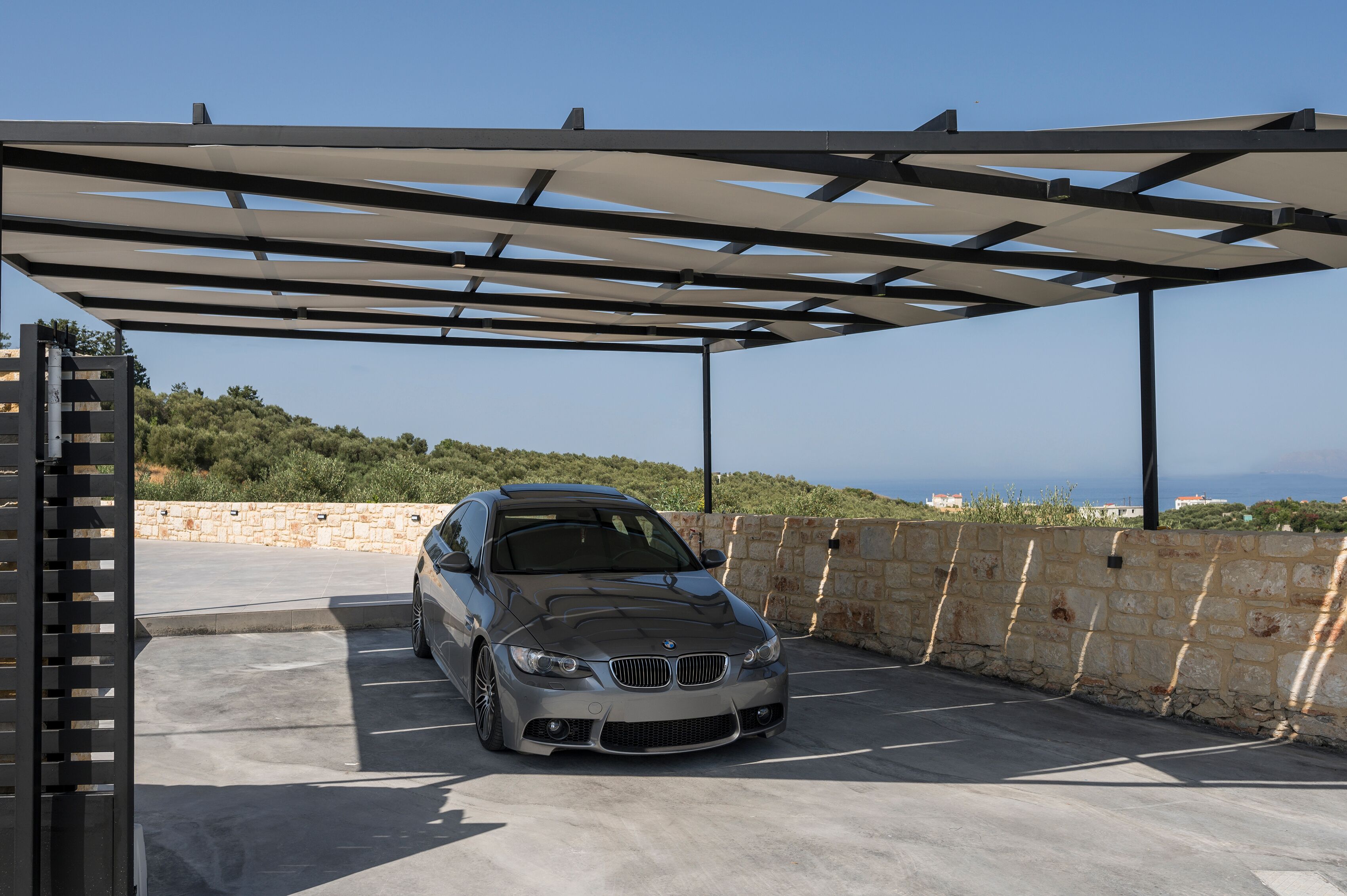 A BMW parked in a covered parking area with a stone wall and a view of the sea in the background.