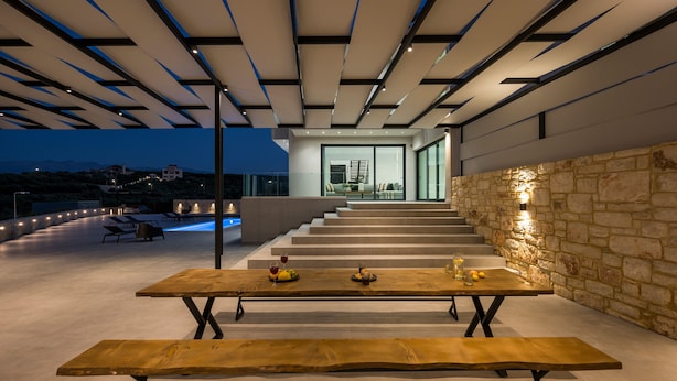 Un espace de restauration extérieur moderne, avec une longue table et des bancs en bois, un mur en pierre et un escalier menant à un niveau supérieur.