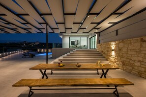 Un espace de restauration extérieur moderne, avec une longue table et des bancs en bois, un mur en pierre et un escalier menant à un niveau supérieur.