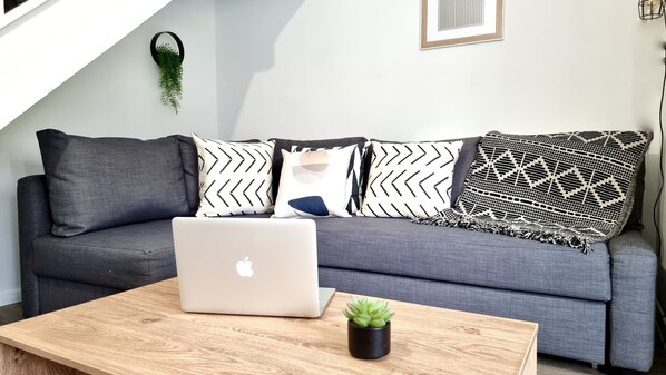 Un sofá gris con cojines estampados, una computadora portátil y una pequeña planta en maceta sobre una mesa de centro de madera.