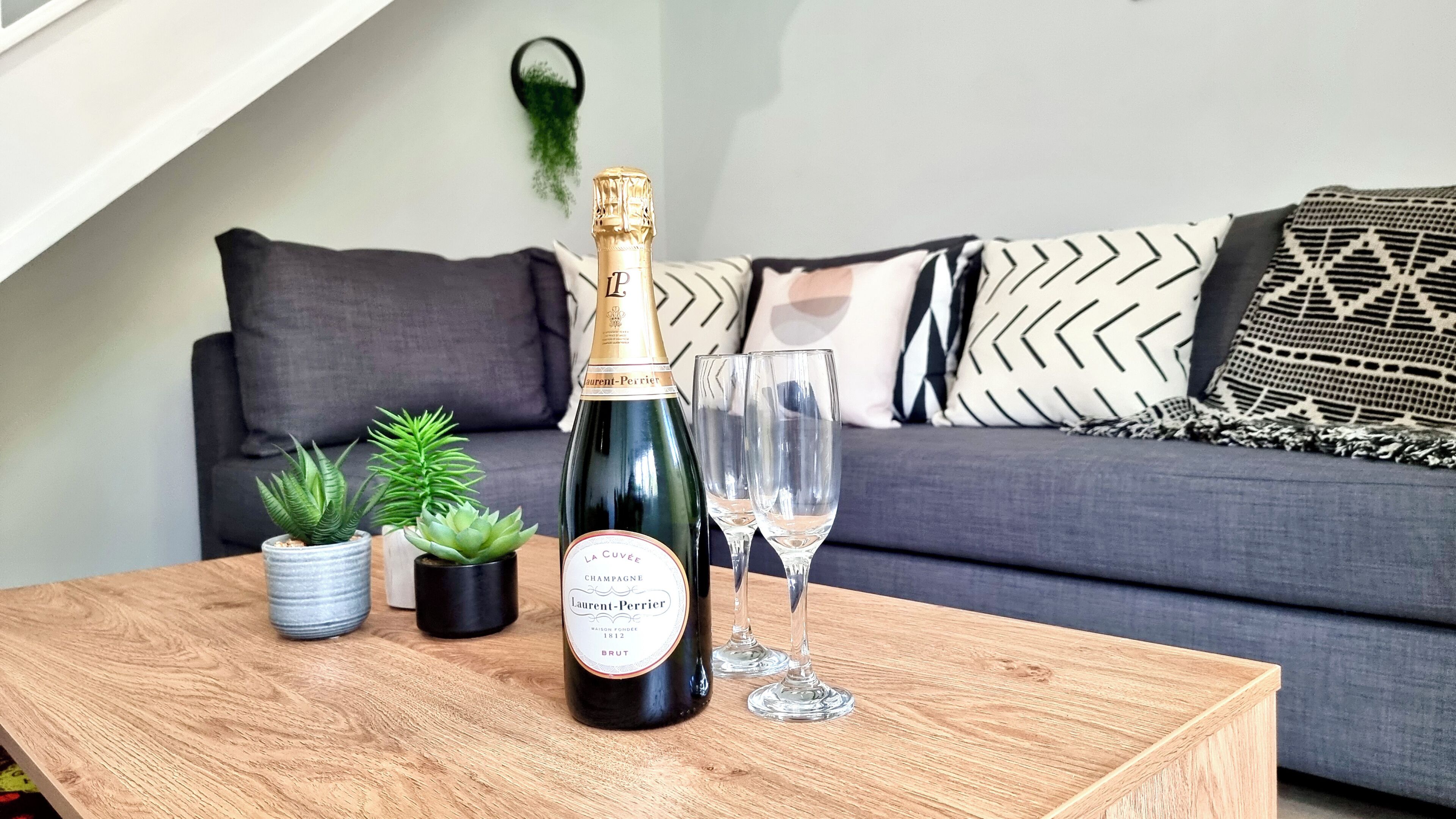Eine Flasche La Cuvee Champagner auf einem Holztisch, daneben zwei Gläser, ein graues Sofa mit gemusterten Kissen und im Hintergrund kleine Topfpflanzen.
