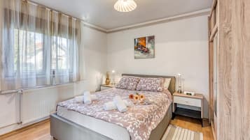 Quarto moderno com uma cama grande, mesa de cabeceira com luminária, radiador e janela com cortinas leves.