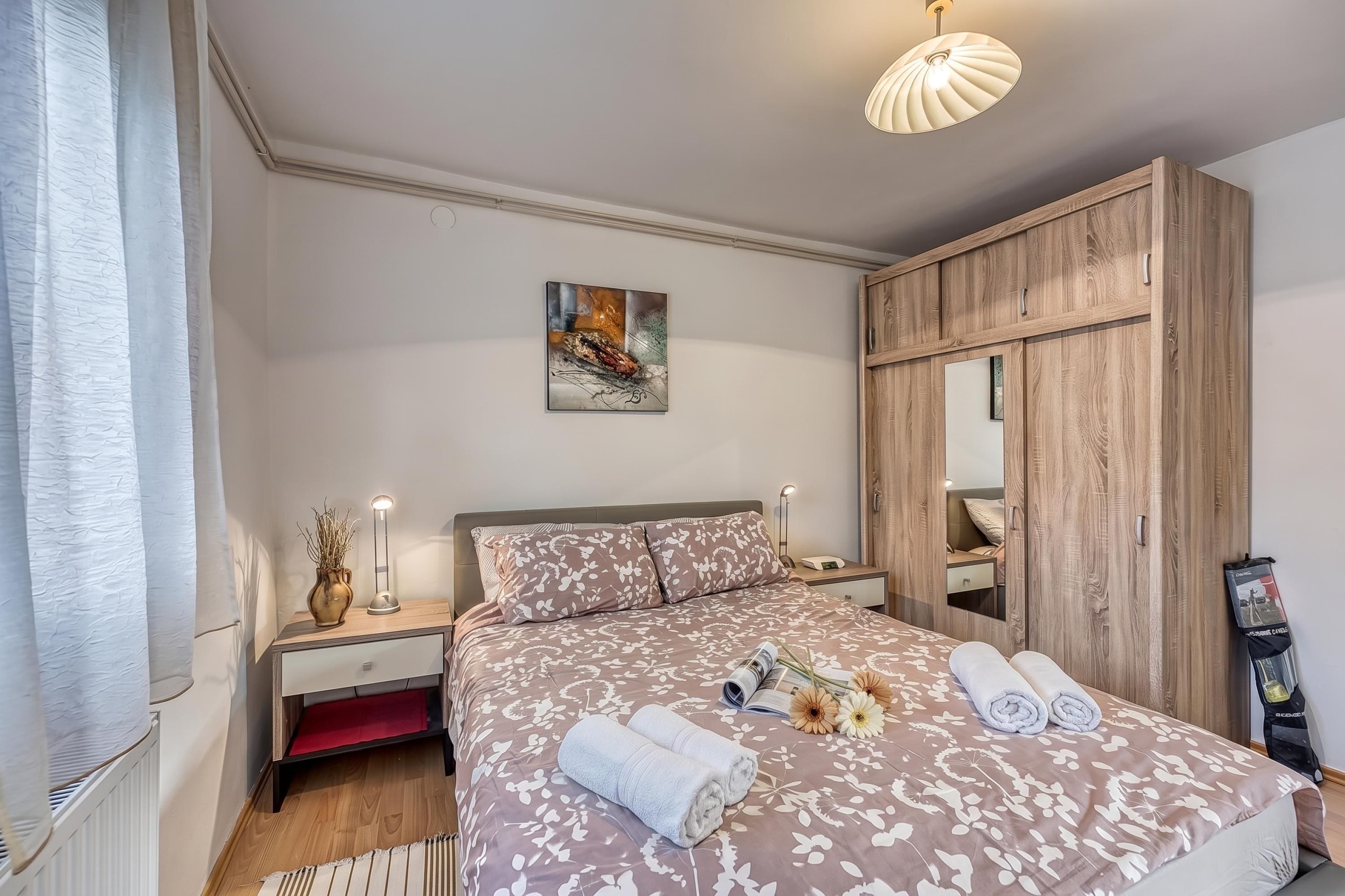 Une chambre à coucher comprenant un lit, des tables de chevet, une commode, un miroir, un luminaire au plafond et une fenêtre avec des rideaux.