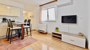A modern kitchen with white cabinets, a dining area with two chairs, a TV mounted on the wall, and a window with curtains. - Lovely apartment few minutes from the beach (Štinjan)