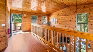 Interior de una cabaña de madera con balcón, pisos de madera y ventanas con persianas.