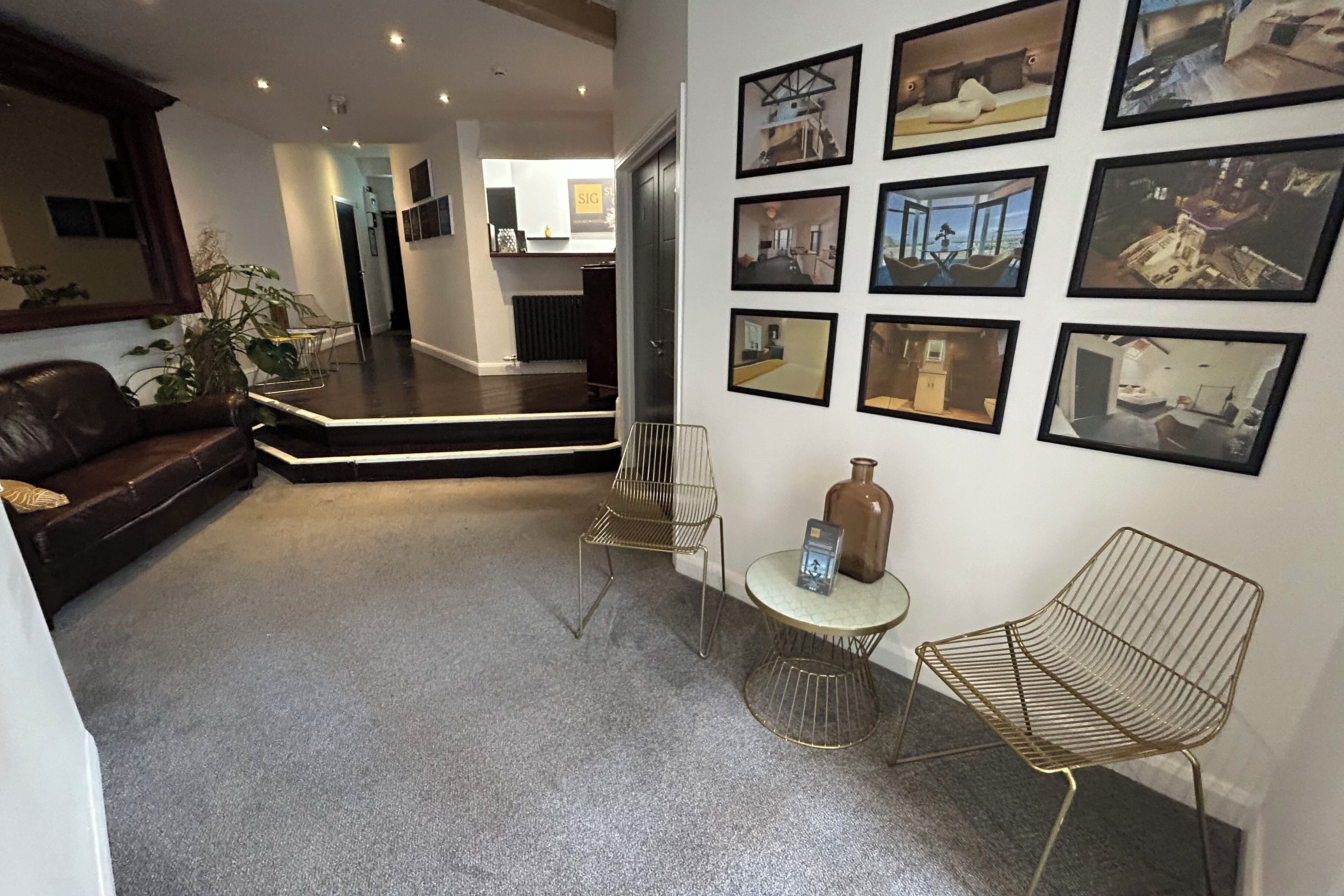 Ein modernes Wohnzimmer mit einem dunkel-lederbezogenen Sofa, einem Couchtisch und einem Stuhl im Drahtgestell. An der Wand hängen gerahmte Bilder und es steht ein Blumentopf bereit.