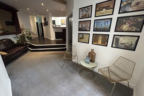 A modern living room with a dark leather sofa, a coffee table, and a wireframe chair. There are framed pictures on the wall and a potted plant. - King Street Serviced Apartments (Lancaster)