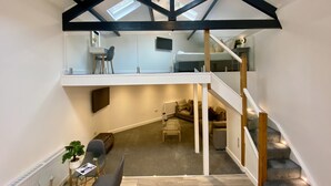 A spacious loft with a high ceiling, exposed beams, a mezzanine level, a staircase, and a cozy seating area. - King Street Serviced Apartments (Lancaster)