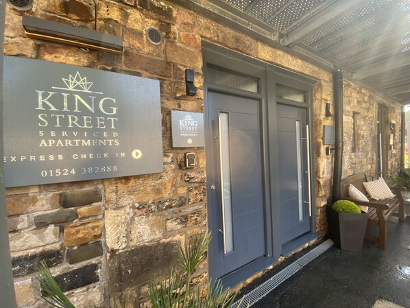 A stone building with a sign for King Street Serviced Apartments, a metal door, and a bench outside. - King Street Serviced Apartments (Lancaster)