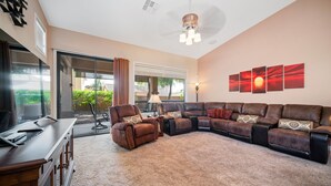 A spacious living room with a large sectional sofa, a small armchair, and a dining table. There is a large window with a view of greenery outside. - Beautiful Single Story Home in Gated Las Sendas Community with Private Hot Tub  (Mesa)