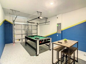 Une table de billard avec un projecteur fixé au plafond, une petite table avec une plante et un mur aux couleurs bleu et jaune.