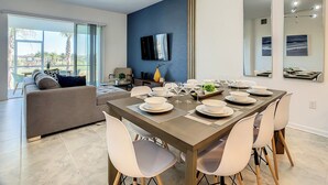 A modern dining area with a large table set for six, a television mounted on the wall, and a balcony with a view of palm trees. - Family Fun: Waterpark & Game Room Condo! (Four Corners)