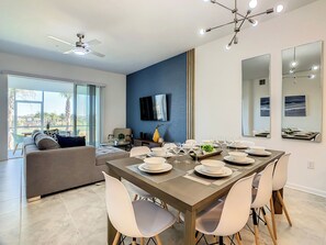 A modern dining area with a large table set for six, a television mounted on the wall, and a balcony with a view of palm trees.