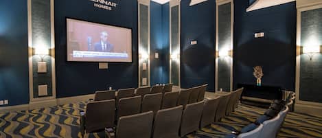 Un salón de conferencias con una pantalla grande mostrando un noticiero, filas de sillas y una alfombra estampada.