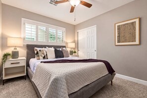 A bedroom with a bed, nightstands, a ceiling fan, a window with blinds, a door, and a framed picture on the wall.