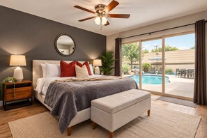 A bedroom with a bed, bedside tables, a ceiling fan, a large window with a view of a swimming pool and outdoor furniture.