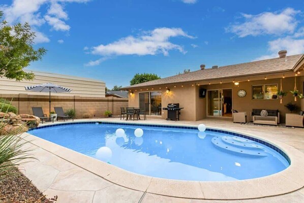Una piscina con borde curvo, mobiliario de exterior y un patio con una parrilla.