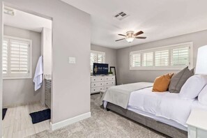 Ein Schlafzimmer mit einem Bett, einem Deckenventilator, einem Schminktisch, einem Nachttisch und einem Fenster mit Jalousien.