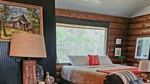 Een rustiek houten interieur met een eettafel, stoelen, een bed met een rood kussen en een groot raam met uitzicht op een bos.