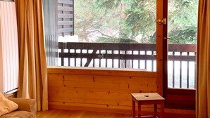 Una habitación con un banco de madera, una mesita y un ventanal con vistas a una terraza de madera y árboles.