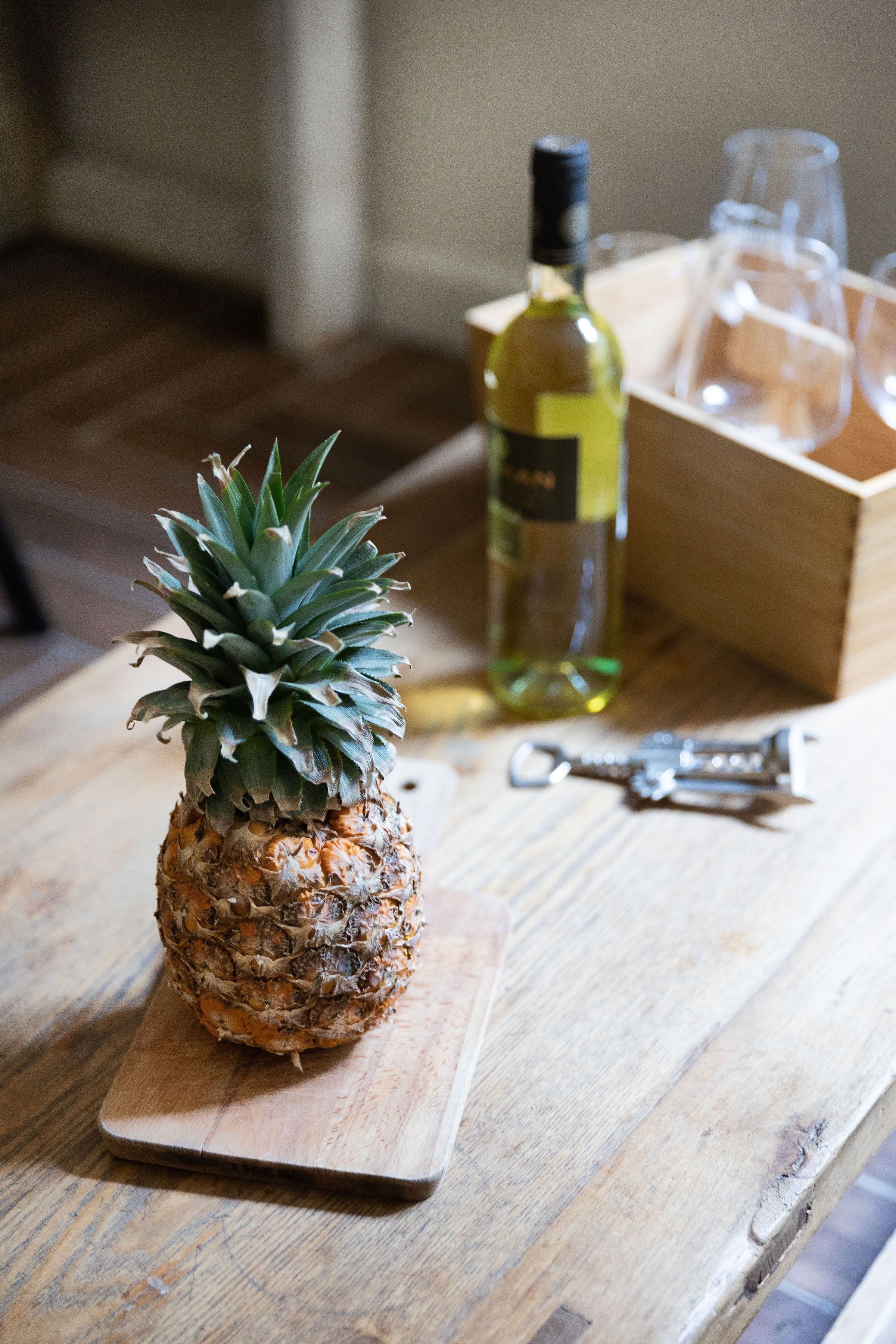 A pineapple on a wooden cutting board, a bottle of wine, and a wine glass on a wooden table.