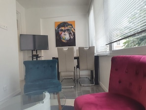 Un salon moderne avec une table basse en verre, un téléviseur à écran plat, deux fauteuils et un tableau aux couleurs vives accroché au mur.