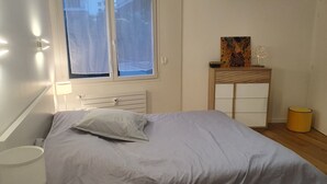 Een slaapkamer met een bed, nachtkastje, lamp, raam, bureau met stoel en een kleine prullenbak.