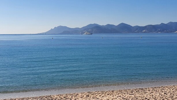 Een strand met helderblauw water, een zandstrand en op de achtergrond bergen.