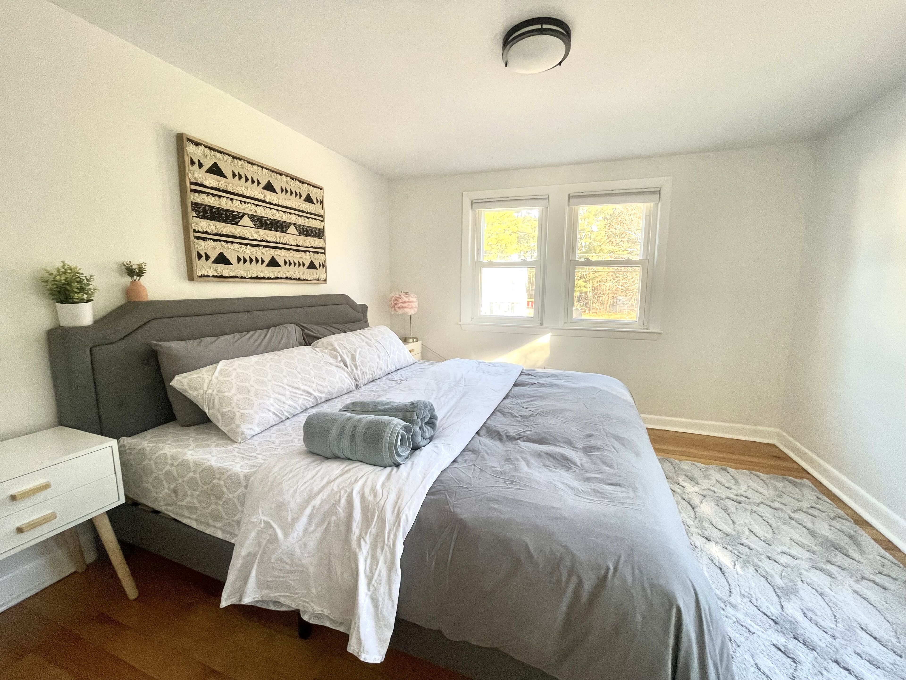 A bedroom with a bed, nightstands, a wall-mounted artwork, and two windows.