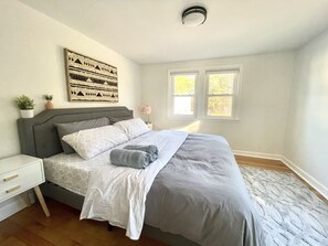 Un dormitorio con cama, mesitas de noche, una obra de arte en la pared y dos ventanas.