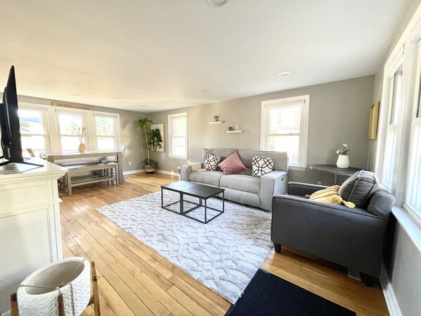A spacious living room with a grey sofa, a coffee table, and a TV. There are windows, a plant, and a dining area with a table and chairs.
