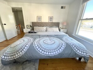 A bedroom with a large bed, a nightstand, a dresser, a window with a view, and a door leading to another room.