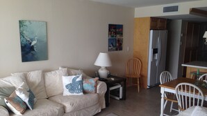 Una sala de estar con un sofá, una mesa, una lámpara y un sillón. Se ve un refrigerador y una mesa de comedor con sillas al fondo.