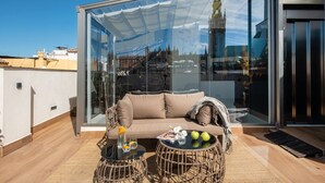 A modern glass-enclosed balcony with a sofa, two wicker tables, and a view of a cityscape including a tower.