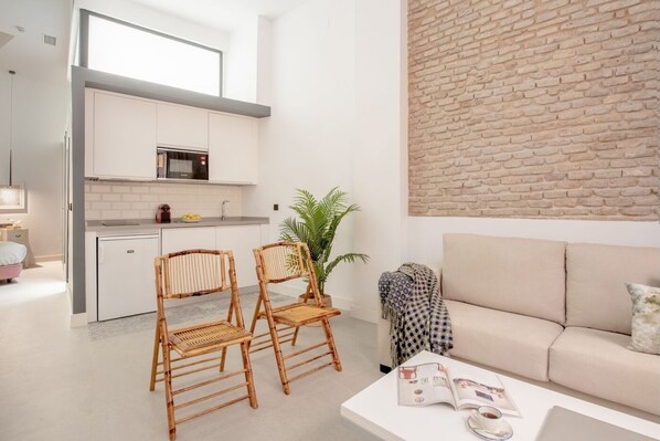 Een modern appartement met een keuken, een zithoek met een bank en twee rieten stoelen, en een metselwerk muur.
