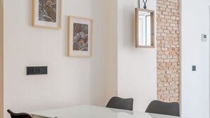 Een moderne eethoek met een witte tafel en zwarte stoelen, kunstwerken op de muur en een accentmuur van baksteen.