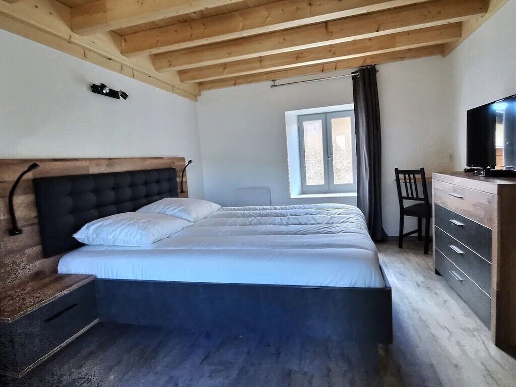 Ein Schlafzimmer mit einem großen Bett, einem Holz-Kopfteil, einem Fenster mit Vorhängen, einem Sessel und einem Fernseher auf einer Kommode.