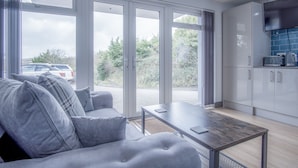 Una sala de estar moderna con un sofá gris, una mesa de centro de madera, y una zona de cocina con armarios blancos y un televisor de pantalla plana.