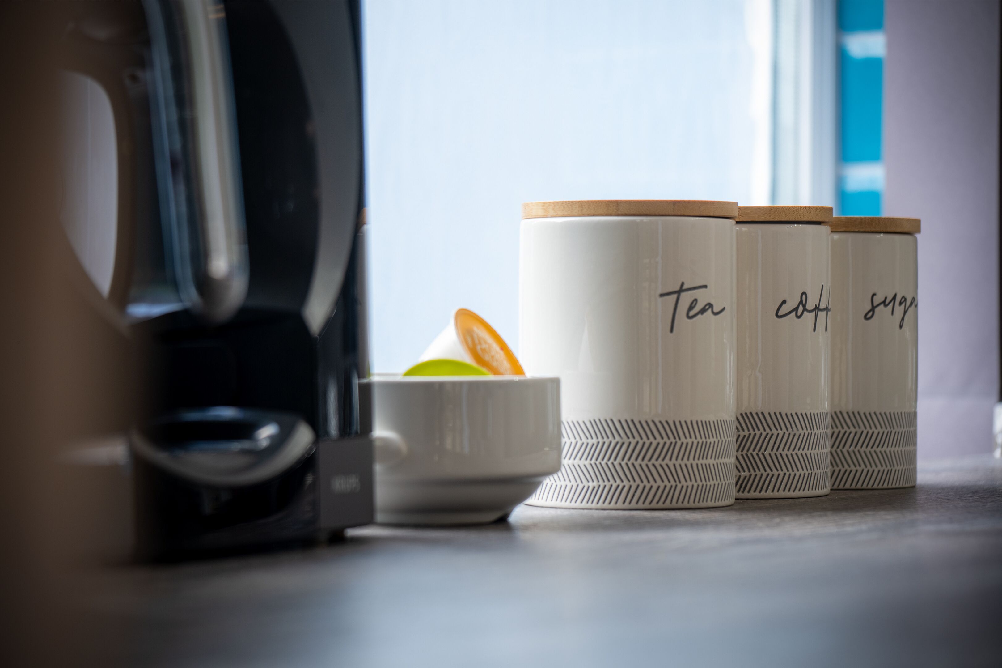 A kitchen area with a coffee maker, a white mug, and three containers labeled "tea," "coffee," and "sugar."