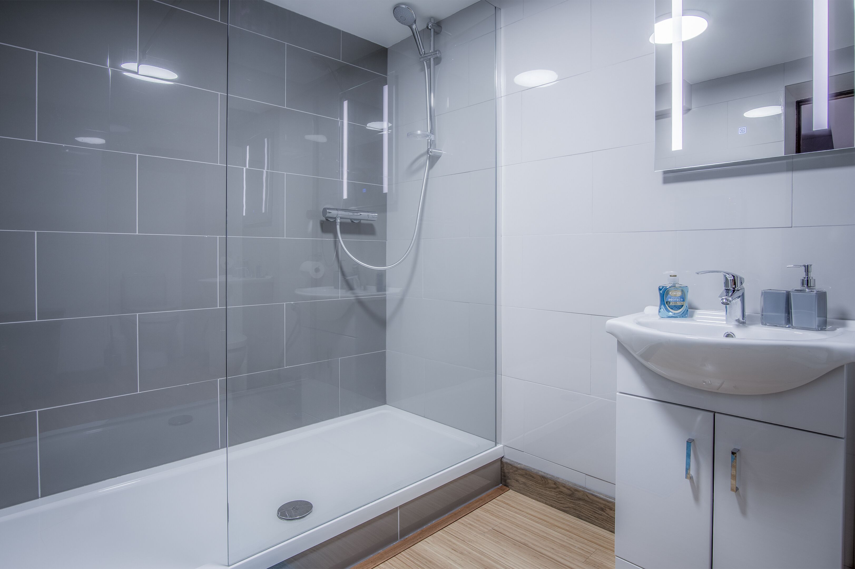 Ein modernes Badezimmer mit Duschkabine aus Glas, einem weißen Waschbecken mit Spiegel darüber und Holzboden.