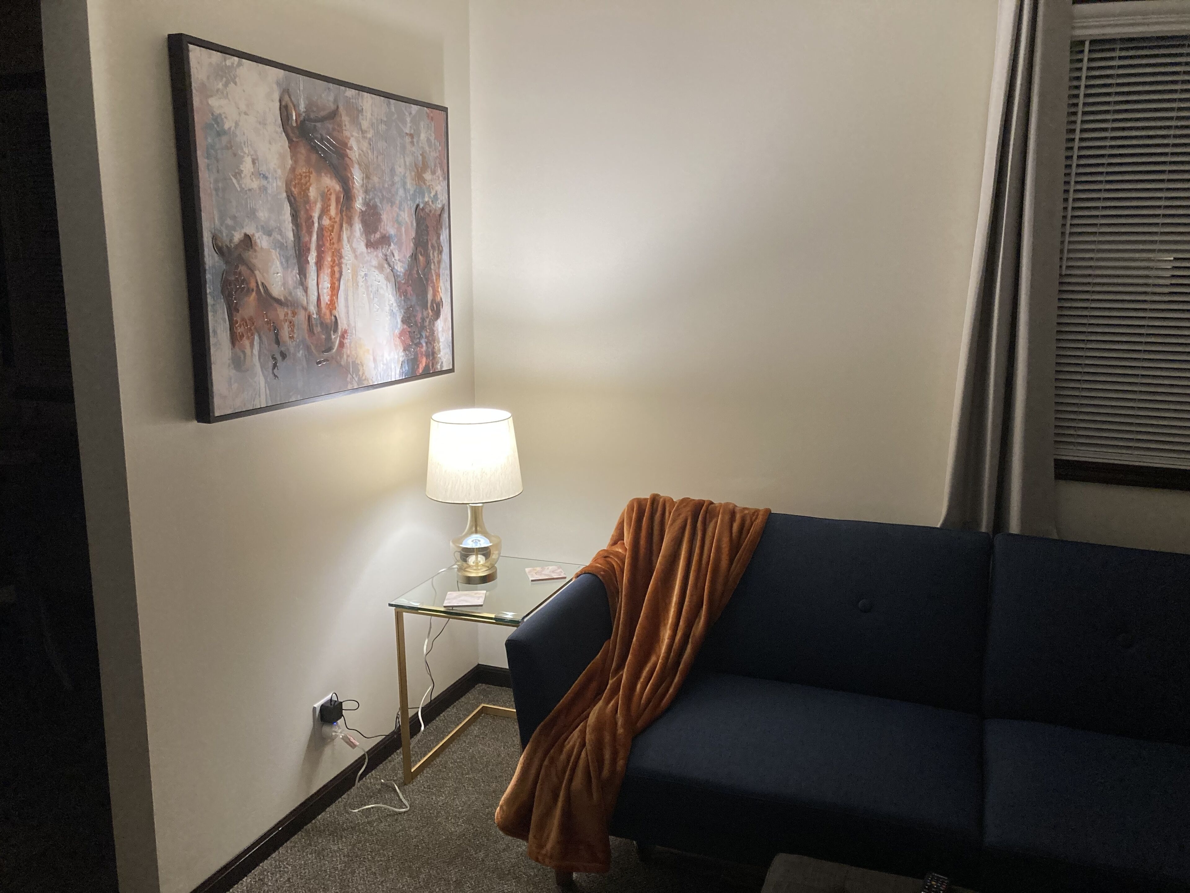 A room with a dark blue sofa, a small glass table with a lamp, a painting on the wall, and a window with blinds.
