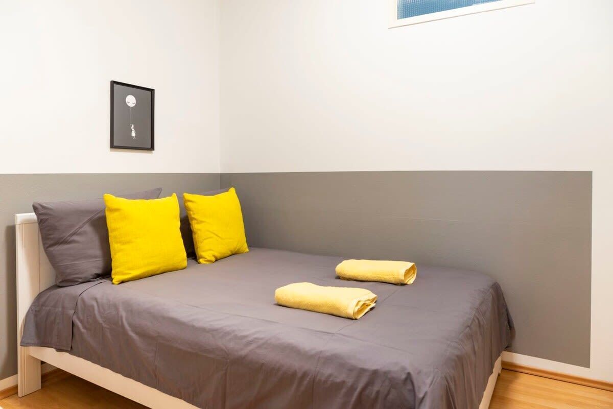 Ein Einzelbett mit grauer Bettwäsche und zwei gelben Kissen, ein gerahmtes Bild an der Wand und zwei aufgerollte Handtücher auf dem Bett.
