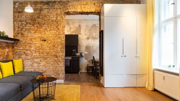 Un espace de vie moderne avec un mur en pierre, un canapé agrémenté de coussins jaunes, une table basse ronde et un coin cuisine visible à travers une arche.