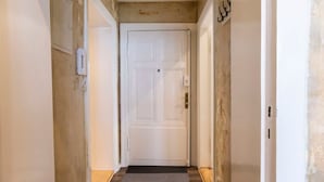 Un couloir étroit, avec une porte blanche au fond, un paillasson et un interrupteur fixé au mur.