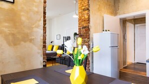 Un coin repas avec une table dressée pour deux personnes, un vase jaune orné de fleurs et un mur en briques.