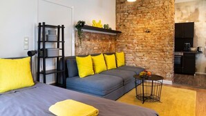 Une chambre moderne avec un canapé gris, un lit recouvert d’une serviette jaune, une petite table ronde sur laquelle se trouvent des fruits, et un coin cuisine visible à travers une porte ouverte.