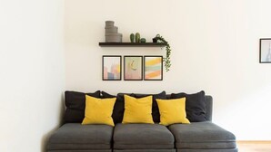 Un canapé gris agrémenté de coussins jaunes et noirs, une petite étagère avec des plantes et un tableau, et trois œuvres d’art encadrées accrochées au mur.