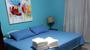 Un letto rifatto con lenzuola blu e asciugamani bianchi, una lampada sul comodino e un quadro astratto colorato appeso al muro.