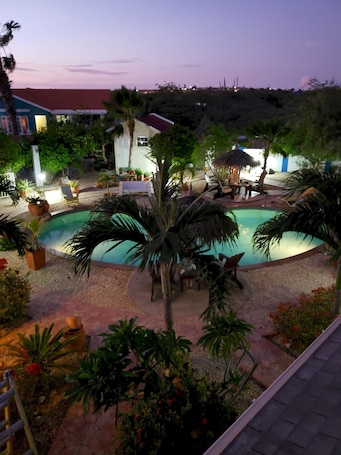 A swimming pool surrounded by palm trees and potted plants, with a few lounge chairs and a thatched-roof structure.